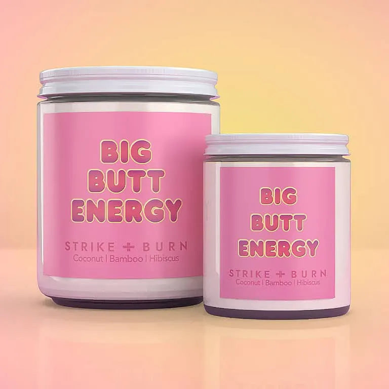 Two jars of 'Big Butt Energy' coconut, bamboo, and hawaiian wax melts by Strike + Burn, shown in a 16oz and 8oz sizes against a soft peach gradient background.